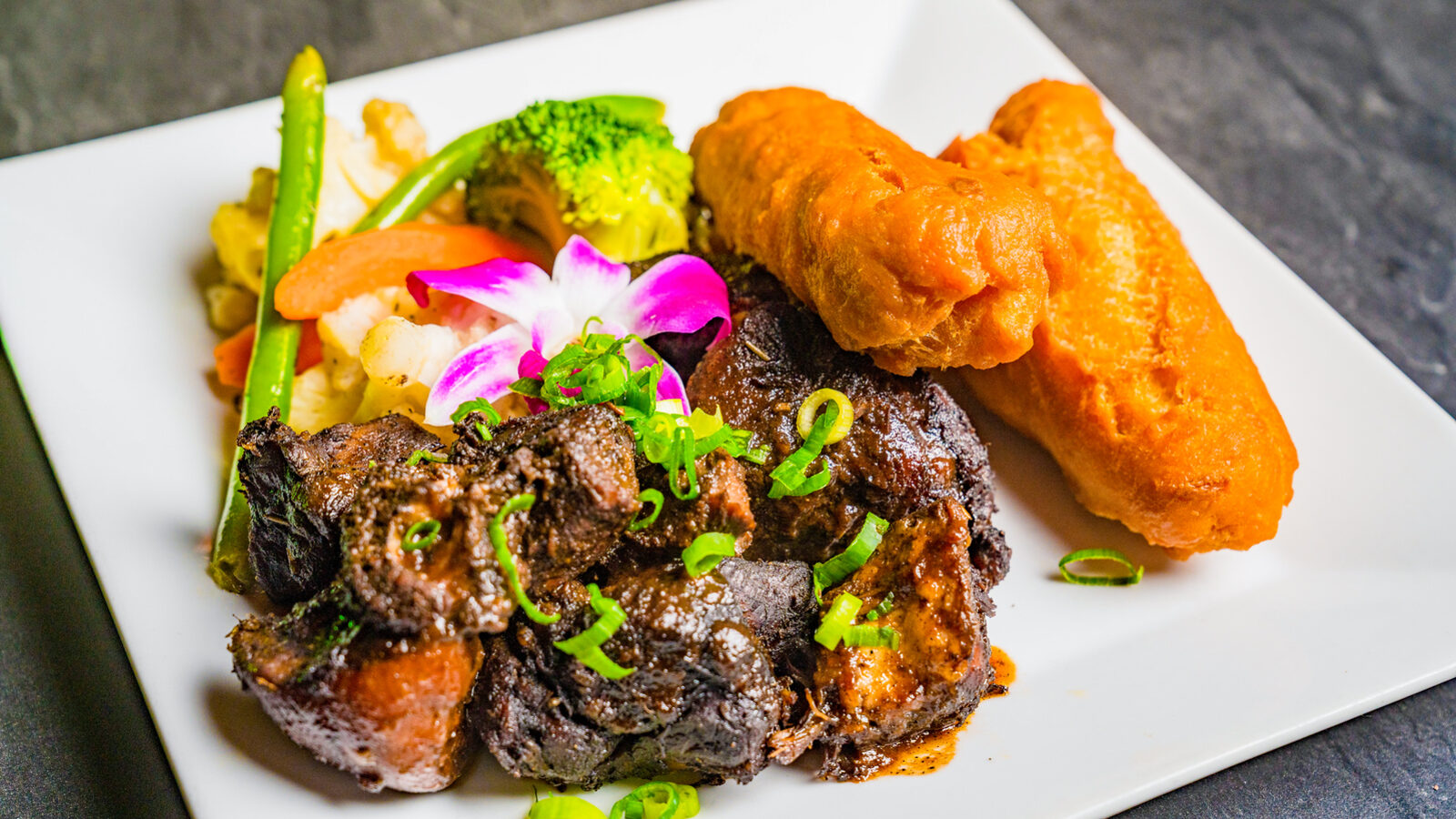 Braised oxtail with fried festival and vegetables on a white plate at Bamboo Walk Caribbean Restaurant
